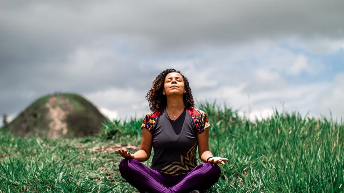 A woman enjoys peaceful meditation in a grassy field under a cloudy sky in Brazil.