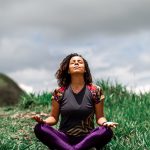 A woman enjoys peaceful meditation in a grassy field under a cloudy sky in Brazil.